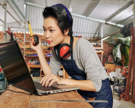 Asian female carpenter with tape measure checking 3D model of furniture piece of tablet computer